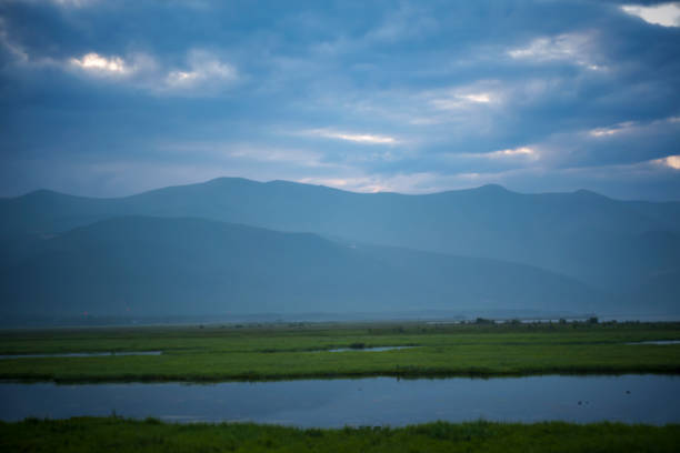 A calm lake with misty mountains at dawn, surrounded by lush greenery under a cloudy sky. The tranquil landscape emphasizes peace and solitude.