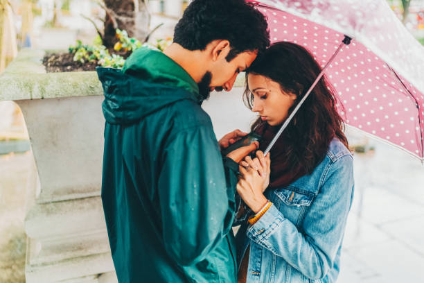 A joyful couple holding an umbrella and capturing a moment together while strolling in a rain-drenched park, reflecting love and companionship in a serene outdoor setting.