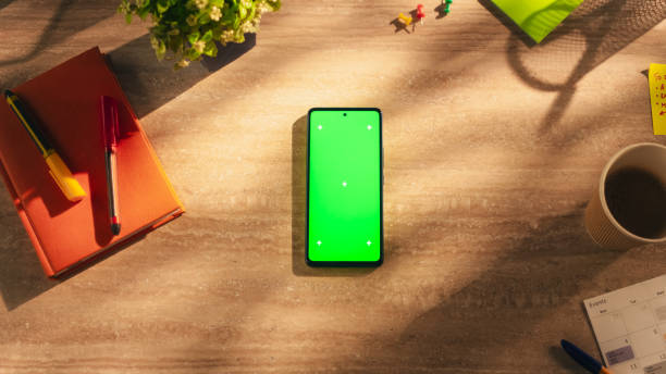 Top Down View of a Smartphone with Mock Up Green Screen Display. Cellphone Device Lying on a Wooden Table in a Creative Office. Template for Online Digital Marketing, Social Media.