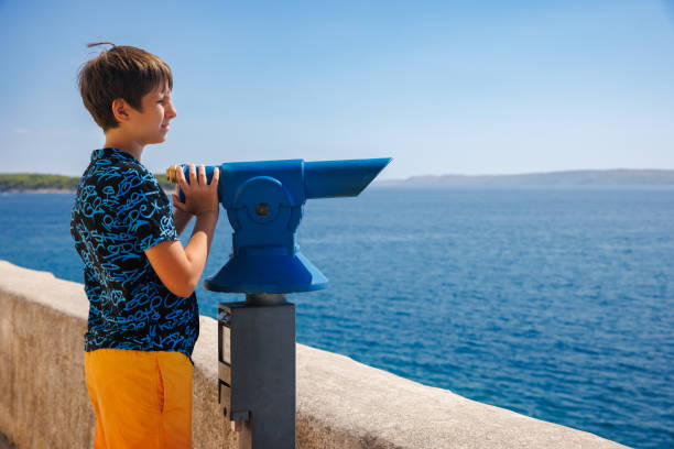 Portrait of cute preadolescent boy standing on the viewpoint by binoculars and looking at the sea, summer vacation on Adriatic Sea