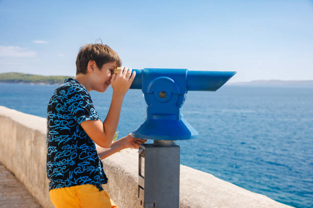 Portrait of cute boy standing on the viewpoint and looking through binoculars at the sea, summer vacation on Adriatic Sea