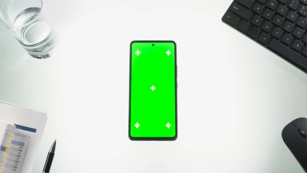 istock Smartphone with Mock Up Green Screen Chromakey Display with Motion Tracker Placeholders. View of a Cellphone Device on a Office Desk with Keyboard, Pen, Documents, Glass Of Water, Computer Mouse 2168768307
