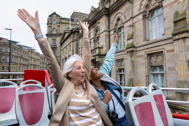 A wide waste-up shot of two joyful senior women throwing their hands up in excitement as they experience an open-top bus tour in the North East of England. They're enthusiastic as they soak in the sights of historic buildings and modern architecture around them. One woman, dressed in a beige coat, and the other in a light blue jacket with a camera around her neck, share a moment of pure delight during their urban adventure. Videos are available similar to this scenario.