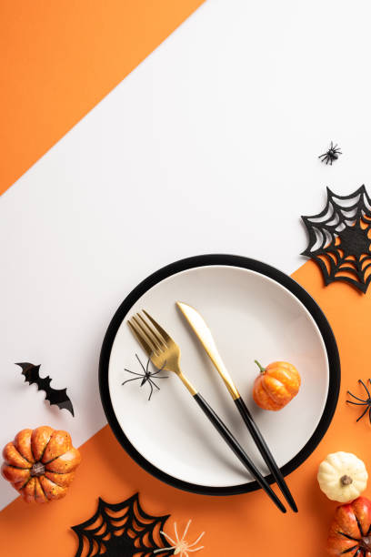 istock Festive Halloween vertical table setting featuring a white plate with gold utensils, miniature pumpkins, spiders, and bat cutouts on an orange background 2168812027