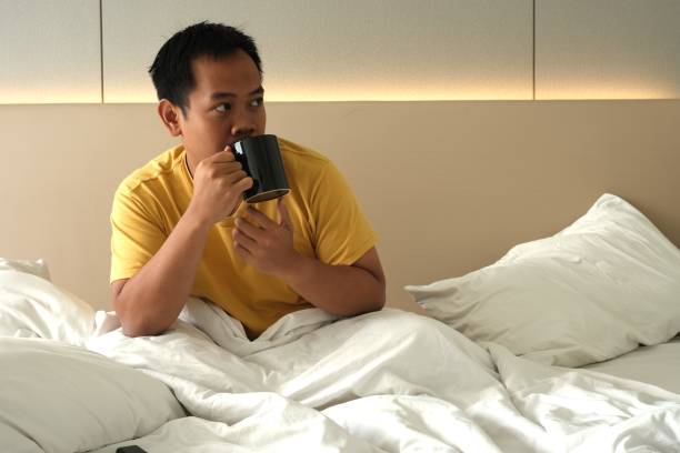 istock Southeast Asian man thoughtfully drinking coffee in bed, surrounded by a soft, well-lit bedroom environment with copy space. 2168814794