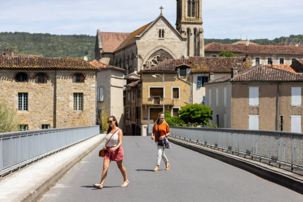 Full-length wide shot of two mature adult females walking across a traffic bridge in the rural French village of Saint Antonin De Noble Val near Toulouse. They are smiling whilst enjoying the views, both wearing casual summer clothing. It is a bright sunny day and in the background historical buildings of the village are visible, including the top of a church.