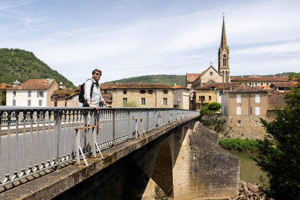 Full-length wide-shot of a young adult male standing on a traffic bridge, admiring views of the surrounding vista, in the rural French village of Saint Antonin De Noble Val near Toulouse. He is leaning against the railing with both hands, wearing casual summer clothing and sunglasses as it is a sunny day. Buildings of the historic village and a church spire are visible beyond the bridge.
