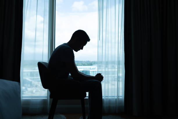 Worried man in boots, jeans and blue shirt is sitting on a top, holding hand on chin, thinking and looking down. Full length studio shot isolated on white.