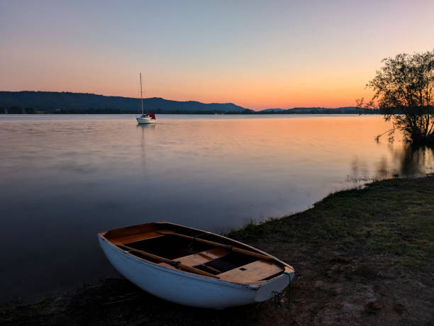 a sailing boat on bodensee at the blue hour with calm smooth water and a rowing boat on the shore