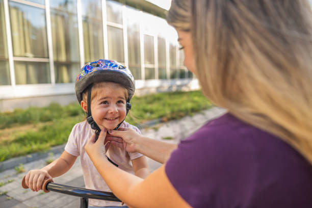 Mother is adjusting the helmet on her daughter's head. She is smiling and getting ready to ride a bike in the park.