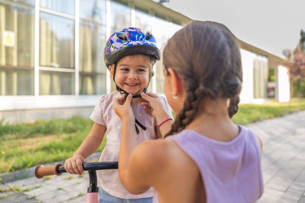 Sister is adjusting the helmet on her younger sister head. She is smiling and getting ready to learning to ride a bike in the park.