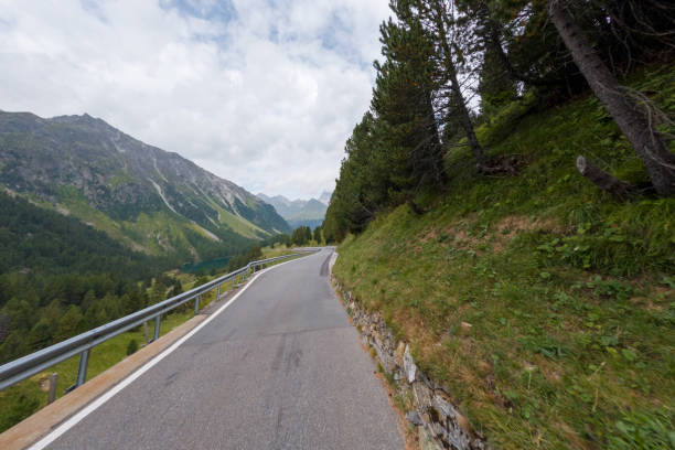 A narrow mountain road winds along a rocky cliffside, with guardrails providing safety and lush green hillsides in the background.