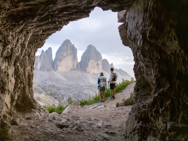 Epic Vista: Hikers Marvel at Tre Cime di Lavaredo's Iconic Peaks from a Dolomites Cave