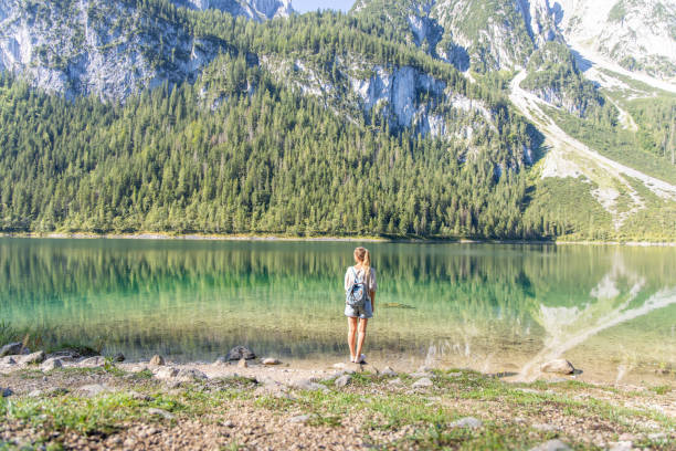 A woman stands at the edge of a crystal-clear mountain lake, gazing at the serene reflection of towering peaks and lush forests.