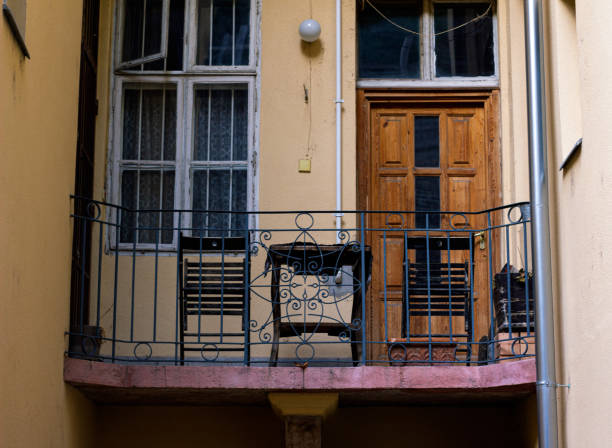 A vintage European balcony featuring a weathered wooden door and an ornate wrought iron railing. The balcony is attached to a yellow building with an old, multi-paned window, partially covered by lace curtains. The architectural details evoke a sense of history, with the chipped paint and aged materials adding character to the scene. The space is small and cozy, typical of urban settings in older European cities.