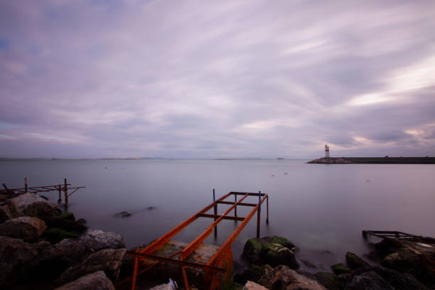 A serene coastal scene captured during the golden hour, showcasing a silhouette of a breakwater with a lighthouse under a breathtaking gradient sky of deep orange and soft blue. A ship with its lights on is visible on the distant horizon, adding a sense of movement and tranquility to the vast sea.