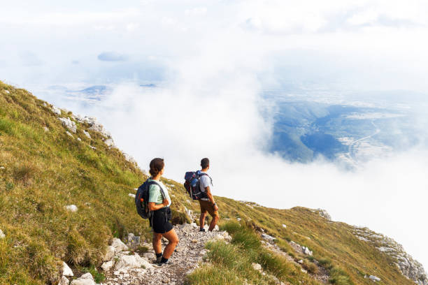 View of a couple on foothpath walking up in the mountain and admiring landscape in the clouds, Gran Sasso National Park, Apennines, Abruzzo, Italy