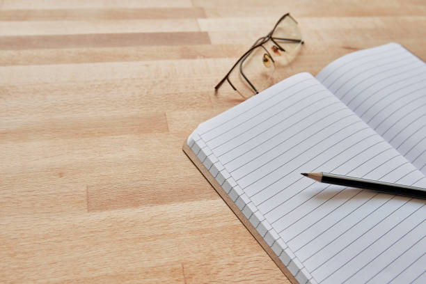 A lined notebook with a pencil resting on its pages is accompanied by a pair of eyeglasses, all placed on a wooden surface. The image offers a significant amount of copy space, making it perfect for themes related to journaling, travel, or writing.