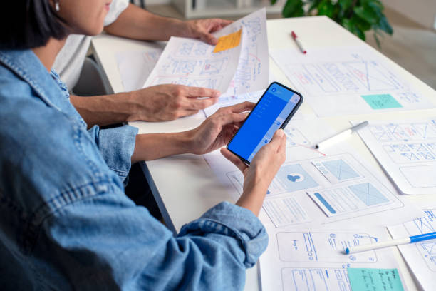 istock UX/UI designer testing prototype on a phone, discussing and brainstorming on wireframes for a website and mobile app prototype, surrounded by sketches of user flow and design tools, in the concept of website and mobile application design concept. 2169040374