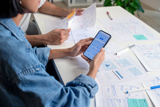 istock UX/UI designer testing prototype on a phone, discussing and brainstorming on wireframes for a website and mobile app prototype, surrounded by sketches of user flow and design tools, in the concept of website and mobile application design concept. 2169040378