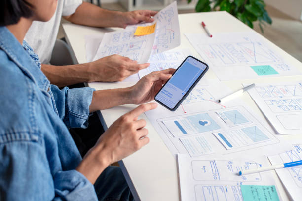 istock UX/UI designer testing prototype on a phone, discussing and brainstorming on wireframes for a website and mobile app prototype, surrounded by sketches of user flow and design tools, in the concept of website and mobile application design concept. 2169040380