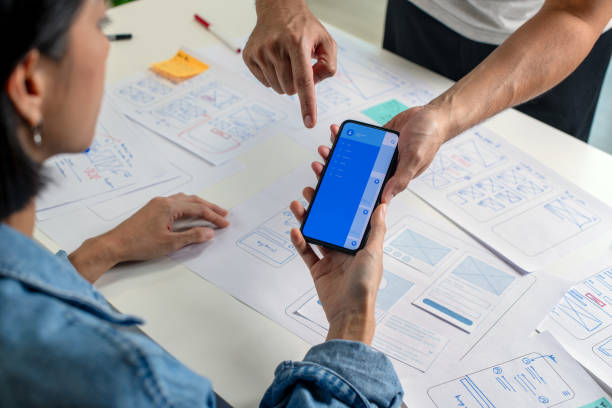 istock UX/UI designer testing prototype on a phone, discussing and brainstorming on wireframes for a website and mobile app prototype, surrounded by sketches of user flow and design tools, in the concept of website and mobile application design concept. 2169040384
