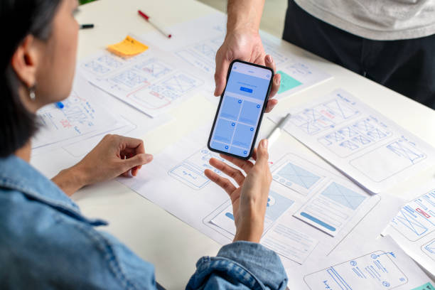 istock UX/UI designer testing prototype on a phone, discussing and brainstorming on wireframes for a website and mobile app prototype, surrounded by sketches of user flow and design tools, in the concept of website and mobile application design concept. 2169040385