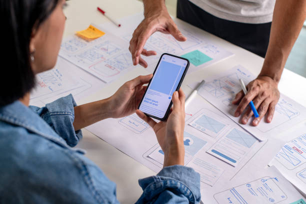 istock UX/UI designer testing prototype on a phone, discussing and brainstorming on wireframes for a website and mobile app prototype, surrounded by sketches of user flow and design tools, in the concept of website and mobile application design concept. 2169040399
