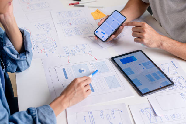 istock UX/UI designer testing prototype on a phone, discussing and brainstorming on wireframes for a website and mobile app prototype, surrounded by sketches of user flow and design tools, in the concept of website and mobile application design concept. 2169040401