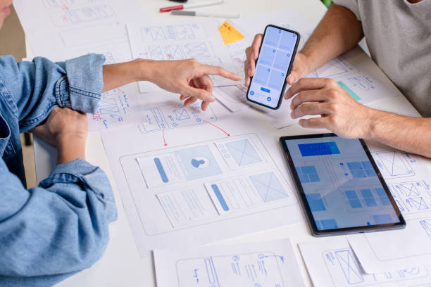 istock UX/UI designer testing prototype on a phone, discussing and brainstorming on wireframes for a website and mobile app prototype, surrounded by sketches of user flow and design tools, in the concept of website and mobile application design concept. 2169040406