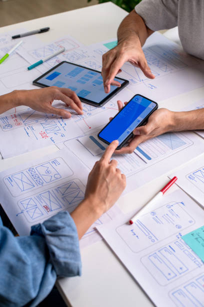 istock UX/UI designer testing prototype on a phone, discussing and brainstorming on wireframes for a website and mobile app prototype, surrounded by sketches of user flow and design tools, in the concept of website and mobile application design concept. 2169040417