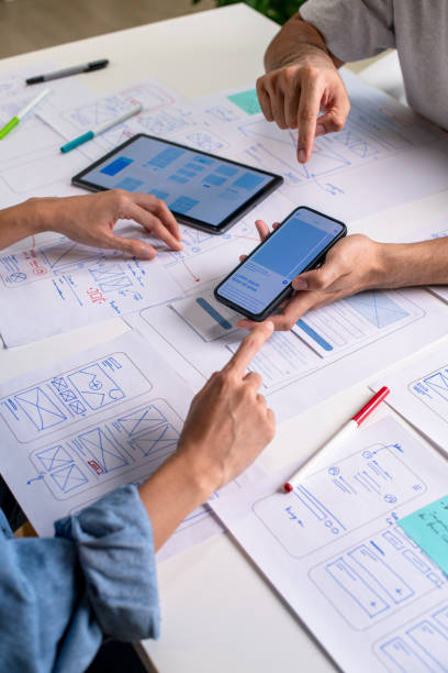 istock UX/UI designer testing prototype on a phone, discussing and brainstorming on wireframes for a website and mobile app prototype, surrounded by sketches of user flow and design tools, in the concept of website and mobile application design concept. 2169040418