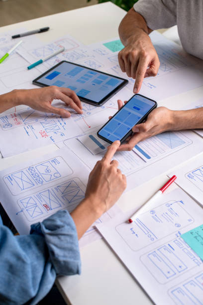 istock UX/UI designer testing prototype on a phone, discussing and brainstorming on wireframes for a website and mobile app prototype, surrounded by sketches of user flow and design tools, in the concept of website and mobile application design concept. 2169040426