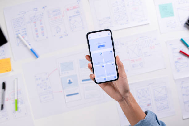 istock UX/UI designer testing prototype on a phone, discussing and brainstorming on wireframes for a website and mobile app prototype, surrounded by sketches of user flow and design tools, in the concept of website and mobile application design concept. 2169040479