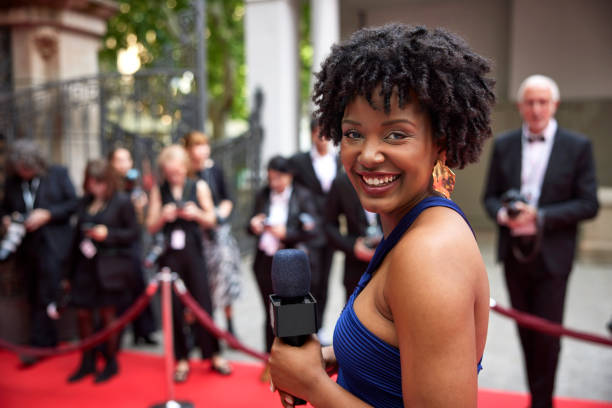 Man in tuxedo arm in arm with woman wearing long gown, smiling and interacting during interview, photographers in background.
