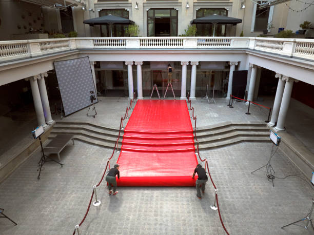 A red carpet entrance lined with gold stanchions and velvet ropes leads to a stage. The carpet is illuminated by a bright spotlight overhead.