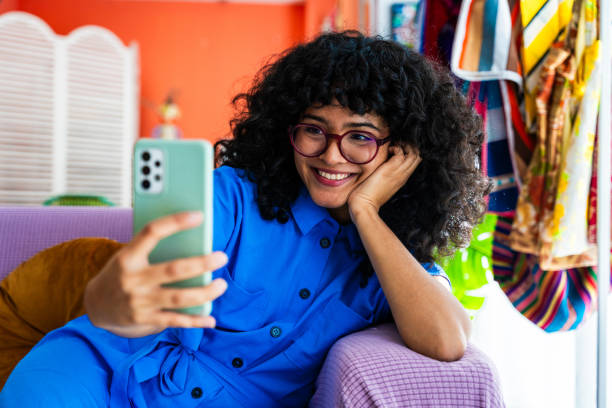 Beautiful young hispanic woman with curly hair portrait at home Beautiful young hispanic woman with curly hair portrait at home - Happy female teenager doing a selfie on the sofa with her smartphone to share on social media multi coloured glasses stock pictures, royalty-free photos & images