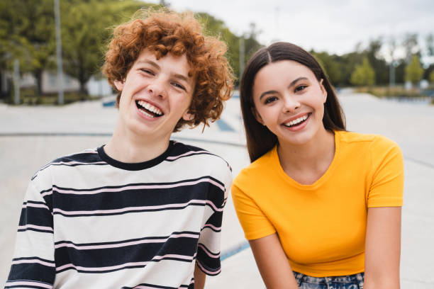 Couple of teenagers in casual clothes sitting laughing have fun in skate park looking at camera Couple of teenagers classmates students high school pupils friends boy girl in casual clothes sitting laughing spend time weekend summer holidays have fun in skate park after lessons looking at camera boy and girl looking at camera stock pictures, royalty-free photos & images