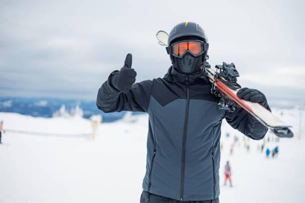 Skier thumbs up, skis on shoulder in a snowy setting. A skier wearing protective gear gives a thumbs up while carrying skis on his shoulder, standing in a snowy outdoor environment, signaling readiness and excitement for the skiing activity. thumbs up for skiing stock pictures, royalty-free photos & images