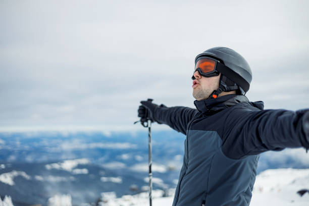 A skier stands on a snowy mountain, arms wide open while holding poles, looking into the distance on a clear day that showcases the expansive natural beauty surrounding them.