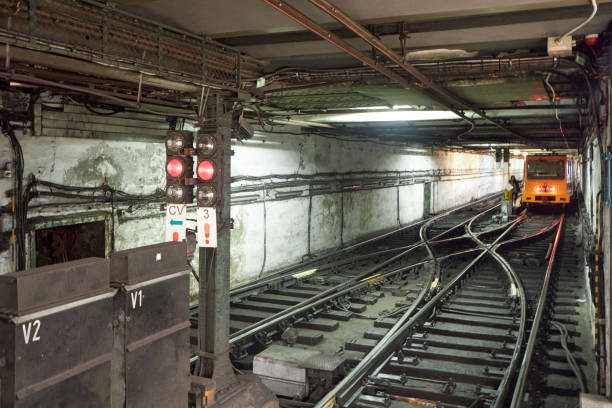 This image shows a section of a metro or train tunnel, featuring multiple sets of railway tracks, signals, and cables running along the walls. The signals are illuminated, indicating that the tracks are in use. A maintenance or service vehicle can be seen in the background, suggesting work being done on the tracks. The condition of the walls and the overhead wiring indicates this is part of an older system, with a utilitarian and industrial feel.\n\nThis scene might be from a maintenance section of an underground metro system, possibly related to the Budapest M1 line or another European metro line known for its historic infrastructure. The junction of the tracks suggests a complex network, often found in older city metro systems.