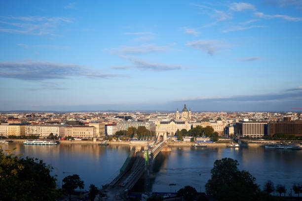 This image offers a stunning panoramic view of a city along a wide river, likely captured during the early morning or late afternoon, as the sunlight softly illuminates the buildings. The river is spanned by a significant bridge, which appears to be the **Chain Bridge** (Széchenyi Lánchíd) in Budapest, one of the most iconic landmarks of the city. The view shows the Pest side of Budapest, with a blend of historic and modern buildings.\n\nIn the background, you can see **St. Stephen's Basilica**, another famous landmark of the city, with its impressive dome rising above the surrounding structures. The Danube River, flowing through the heart of Budapest, reflects the calmness of the day, and the skyline offers a mixture of architectural styles, highlighting the city's rich history.\n\nThis view is likely taken from the Buda side, perhaps near the Buda Castle or Gellért Hill, providing a picturesque overview of the city.