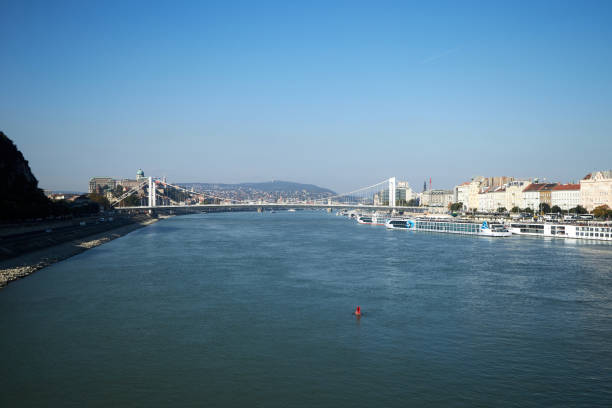 This serene photograph captures the expansive beauty of the Danube River, flowing gracefully through a vibrant urban landscape. The image is taken from a vantage point that showcases the river’s wide expanse, reflecting the clear blue sky above. The calm water contrasts beautifully with the rocky riverbank on the left, adding a touch of natural ruggedness to the scene.