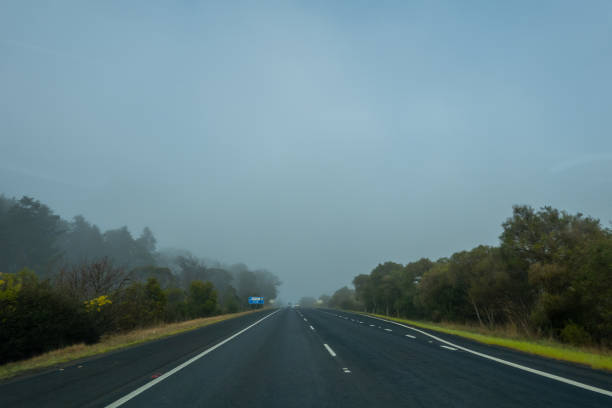 The scene captures the view from inside a car driving down a motorway blanketed by mist and fog, significantly reducing visibility. The headlights of vehicles ahead are faintly visible through the thick fog, while lane markers and road signs appear blurred and distant.