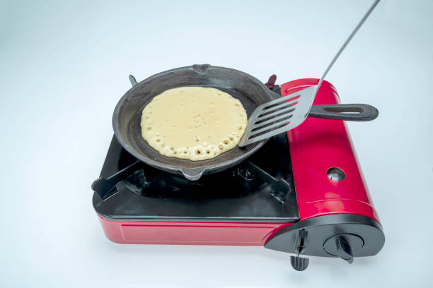 A pancake is being cooked on a red stove top. The pancake is golden brown and is being flipped with a spatula. The stove top is a small, portable one that can be used for cooking on the go