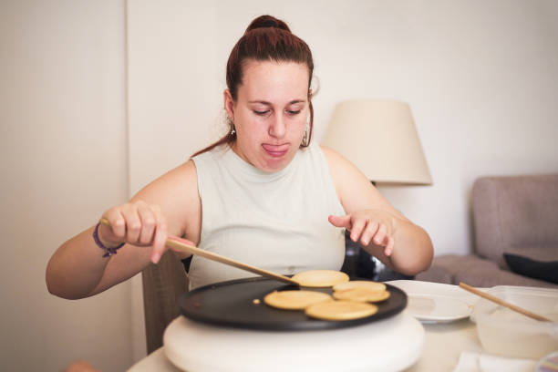 A disabled woman is cooking pancakes on a pan. She is using a wooden spatula to flip the pancakes