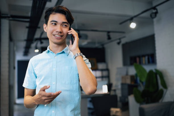 istock Young businessman wearing a smart casual shirt smiling while using a smartphone in a modern office environment with plants and books. 2169216307