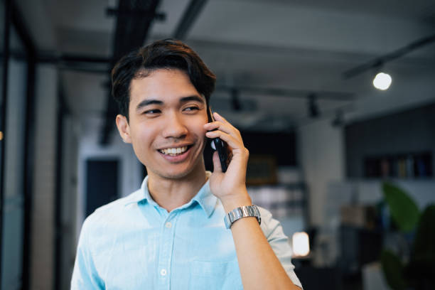 istock Smiling young businessman talking on a mobile phone in a modern office setting, wearing a light blue shirt and a silver wristwatch. 2169216731