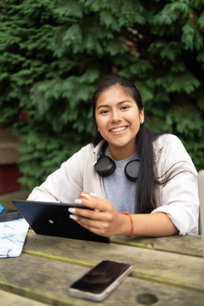 istock Young hispanic female student smiling while using a tablet to study at an outdoor campus table 2169219380