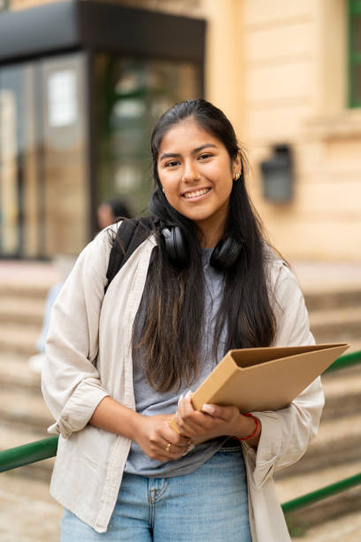 istock Young woman is smiling while standing outside on a campus holding a folder and wearing headphones around her neck 2169219414